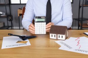 Bankrupt businessman with calculator and house model at table in office