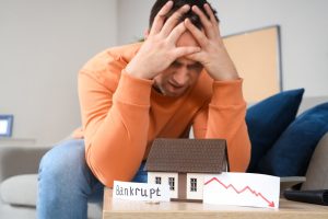 Paper with word BANKRUPT, downward arrow and house model on table of stressed man at home